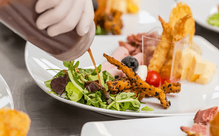Chef drizzling dressing over a fresh mixed green salad with cherry tomatoes and cheese on a white plate.
