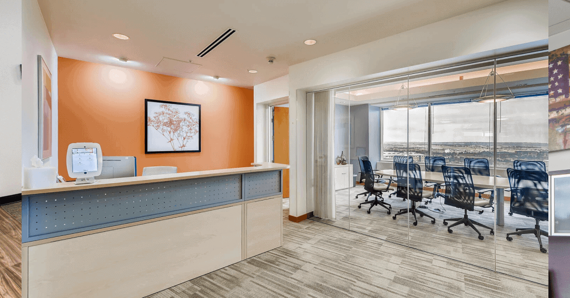 Modern office reception area with a front desk, orange accent wall, and large glass conference room with chairs and city view.